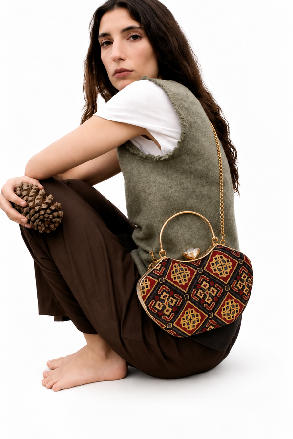 Kaali Adaa curved Mashru silk handbag in a black, red and mustard diamond print, on its gold-tone chain with a seated model in a green knit vest