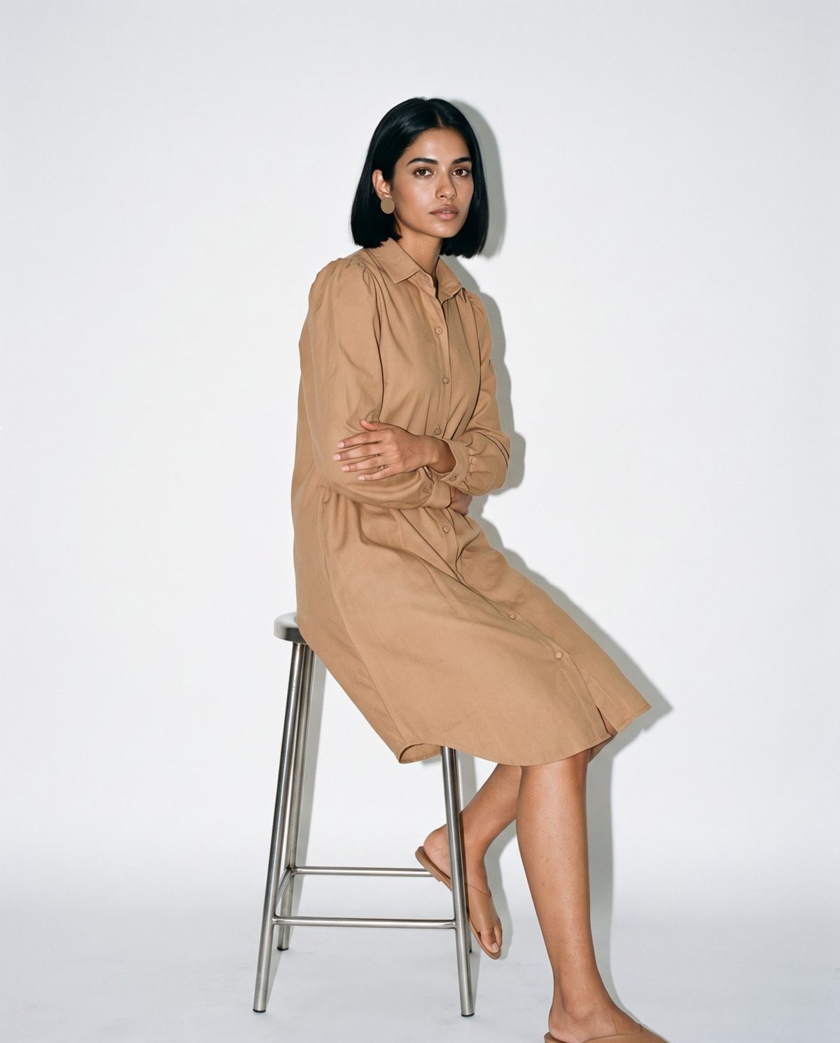 Ujaas cover: a model seated in the Ibrik camel shirt dress and Ibrik Accents earrings in a white studio