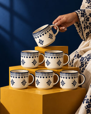 Six Saanjh cups with a navy floral print stacked on yellow plinths, one lifted by hand against a deep blue wall
