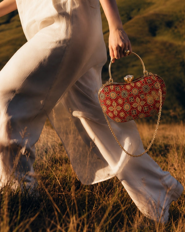 Rang Mahal red Mashru silk handbag carried by its arched top handle, model walking in ivory trousers through tall grass