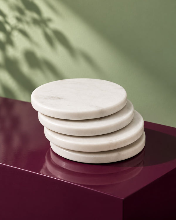 Table Companions, Hand-Carved Marble Coasters (Set of 4) four coasters stacked on a maroon plinth in dappled light (AI-generated styling image)