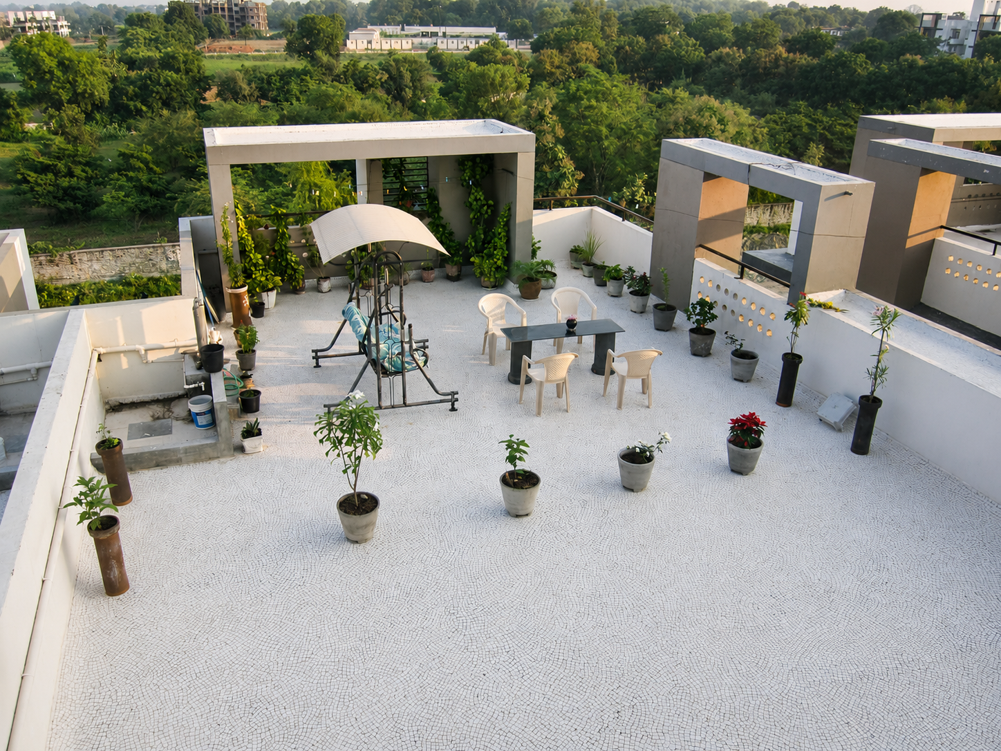 Rooftop terrace at dawn in an Indian home: a canopied jhula swing, a hand-tended potted garden of tulsi, hibiscus and poinsettia, and chairs around a table, with green fields beyond.