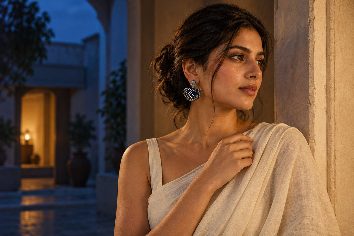 AI-generated editorial: Blue Chandbali fabric earrings with white polka dots and silver ghungroo, worn with an ivory drape at dusk.