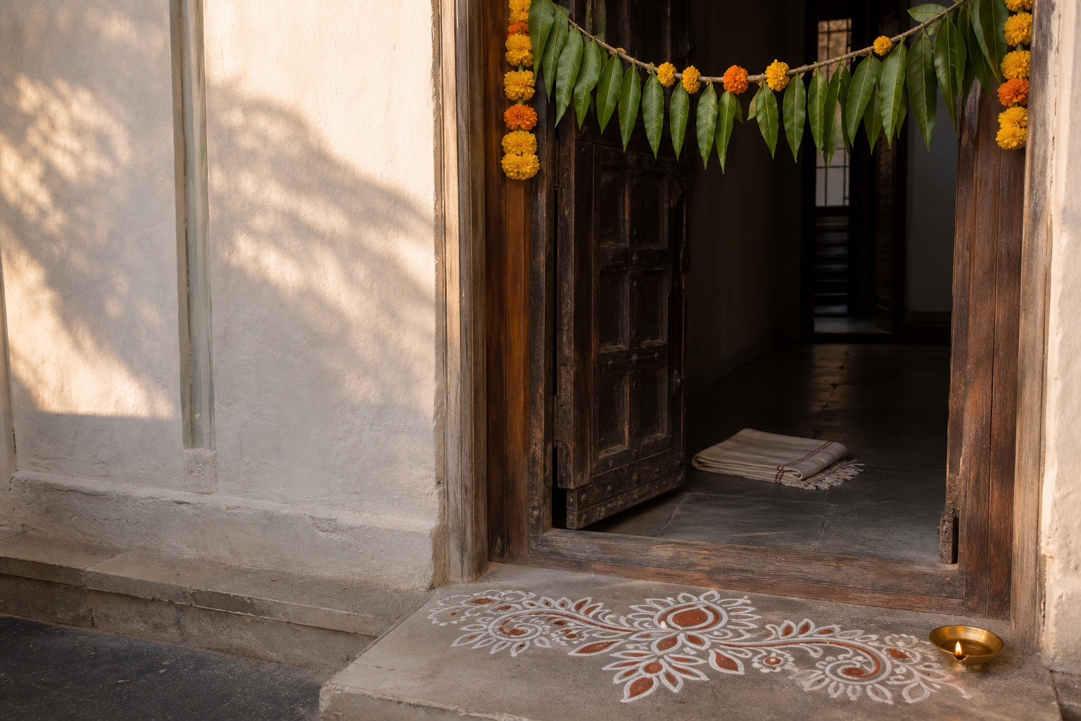 An Indian doorway at dawn with a fresh white-and-rust rangoli, mango-leaf toran and brass diya in warm natural light