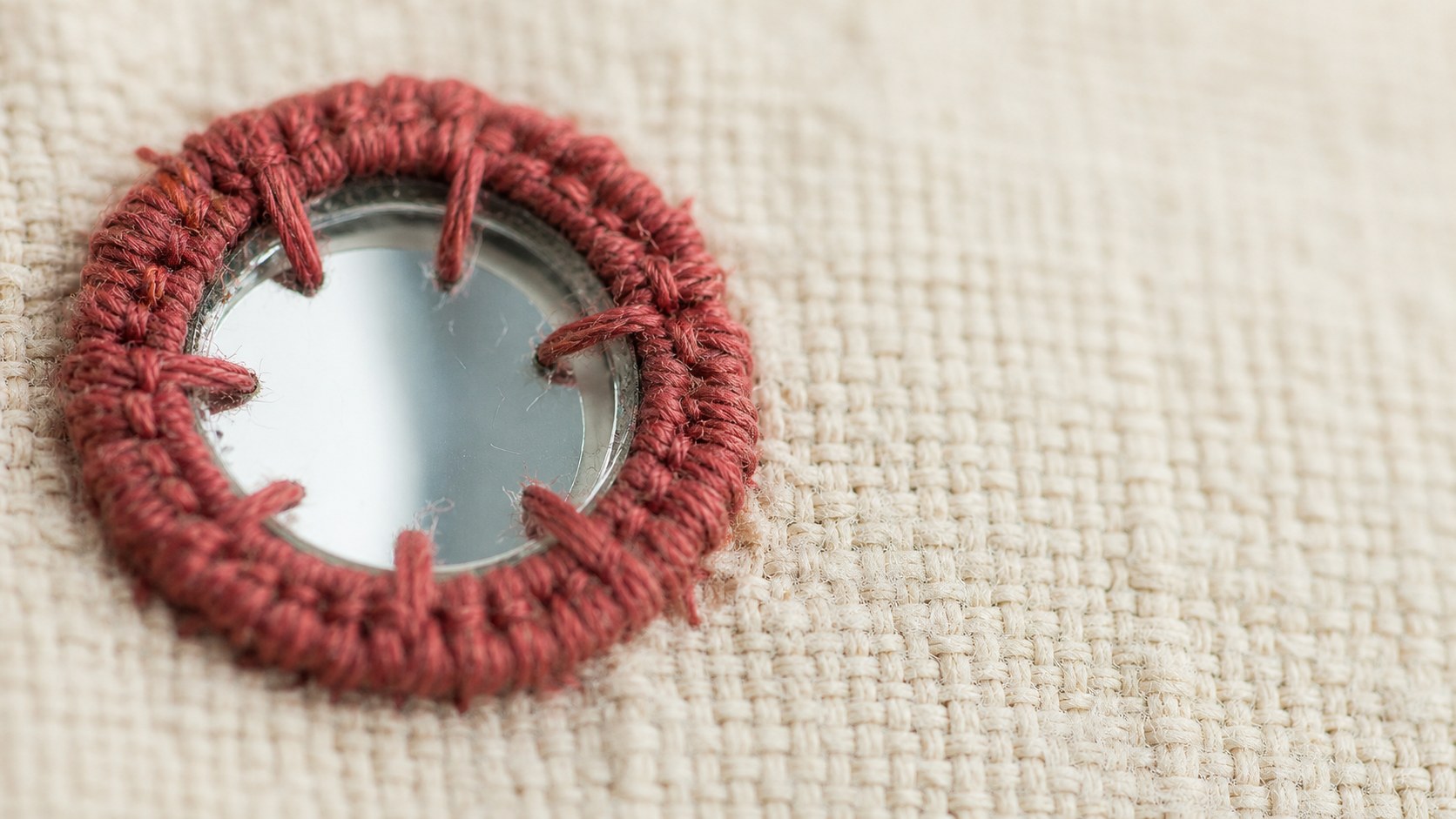 Macro of a mirror held to cloth by a collar of rust-red thread crossing its rim — hand-set abhla mirror work, House of Falguni