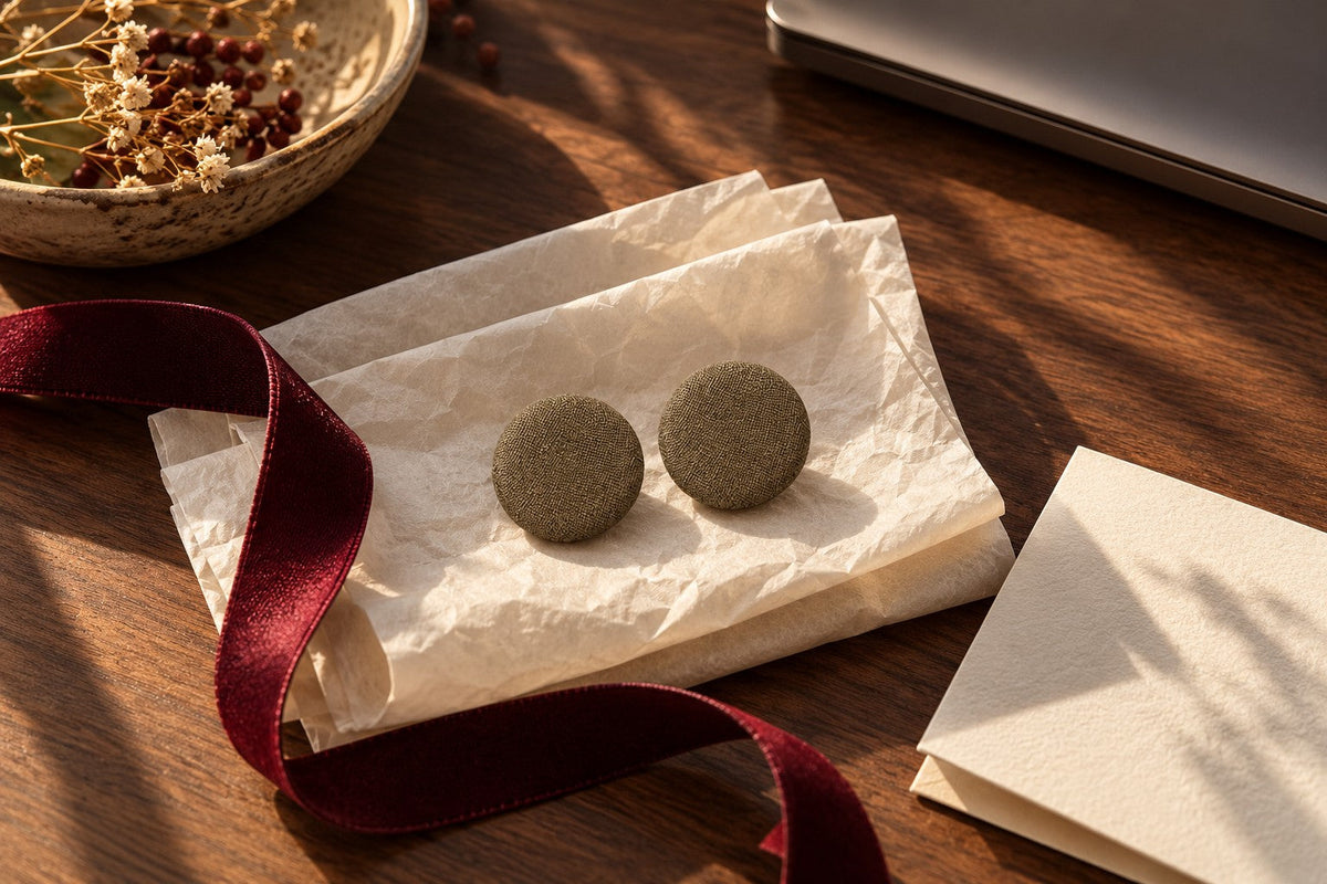 A pair of plain round Ibrik fabric studs beside tissue and an oxblood ribbon on a walnut desk.