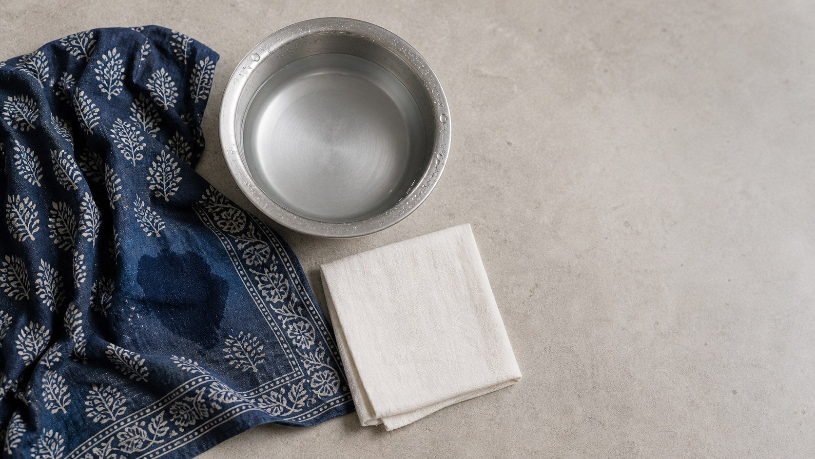 Block-printed indigo cloth with a fresh spill beside a bowl of cold water and a folded white blotting cloth — how to remove stains from ethnic wear, House of Falguni