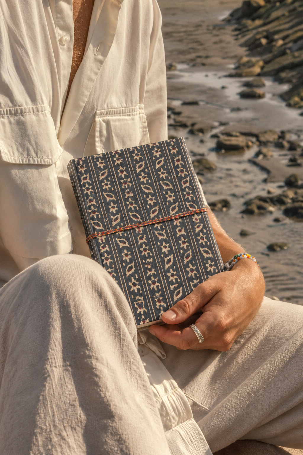 Indigo Ajrakh block print fabric diary resting on the knee of someone in cream linen, seated on a rocky shore