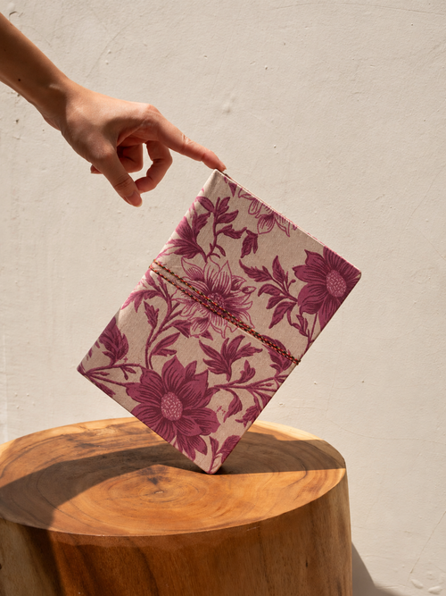 Blush Garden block print fabric diary balanced on a wooden stump stool, a fingertip steadying it against a cream wall