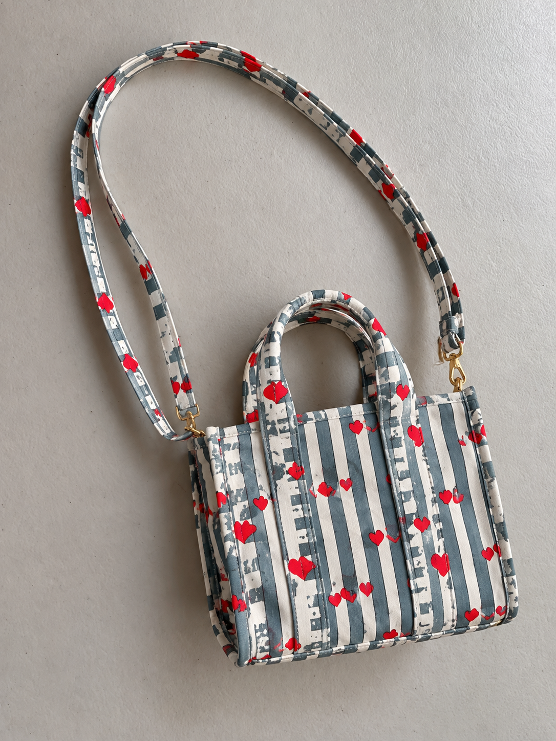 Jal quilted mini tote in grey and white stripes with small red hearts, lying flat with its long strap attached