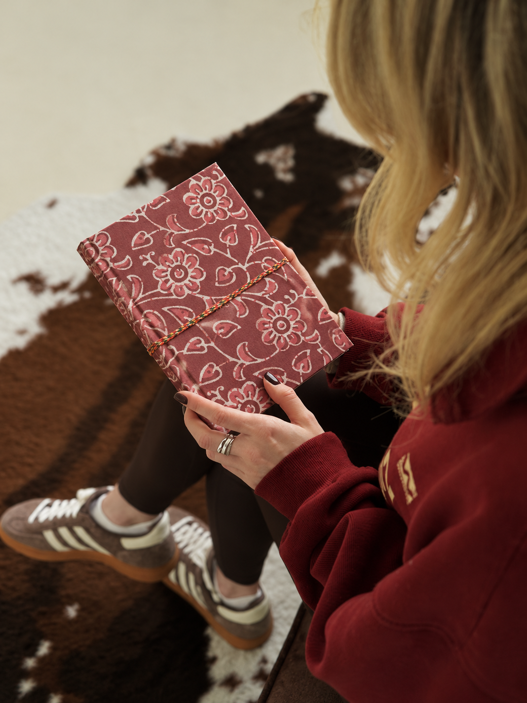 Block print fabric diary with outlined flowers and heart-shaped leaves, read on a cowhide rug by a woman in a burgundy sweatshirt
