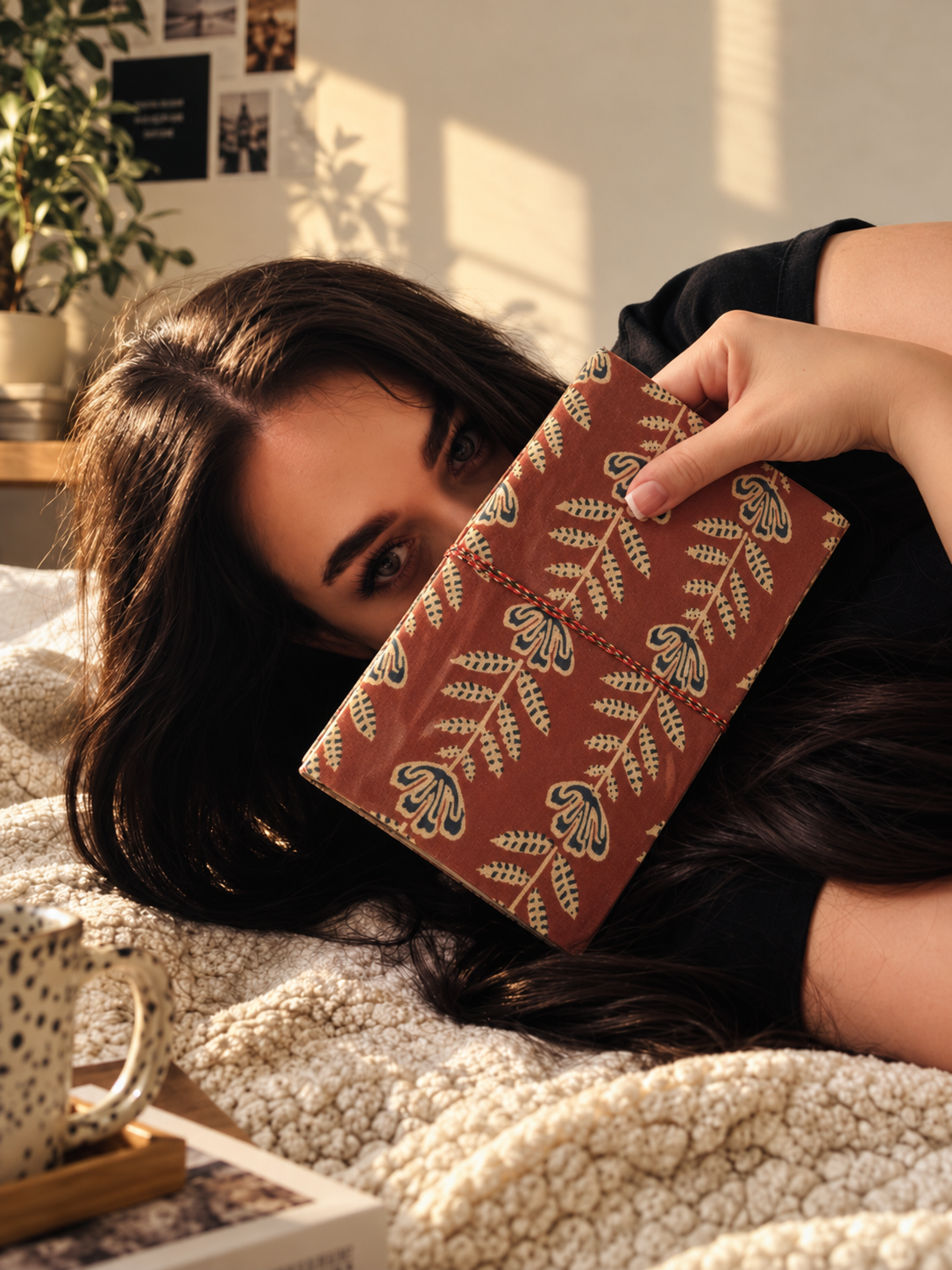 Crimson Jaipur block print fabric diary held across the face by a woman lying on a cream boucle throw in afternoon sun