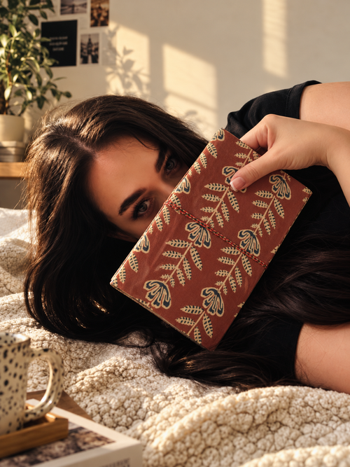 Crimson Jaipur block print fabric diary held across the face by a woman lying on a cream boucle throw in afternoon sun
