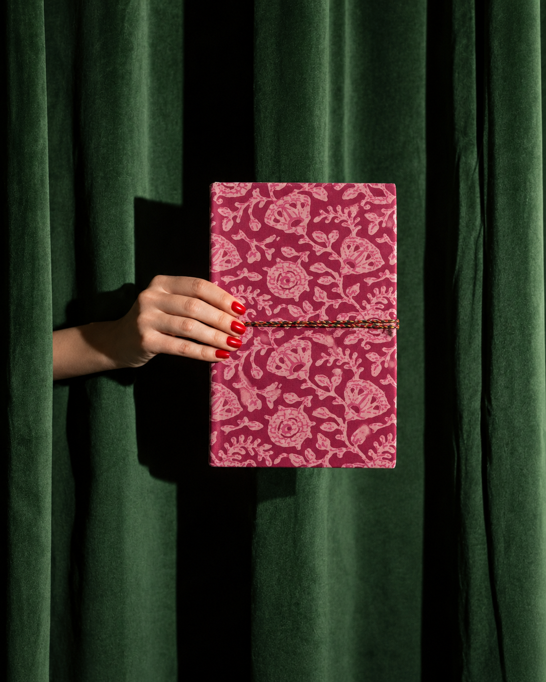 Pink block print fabric diary with dense pale florals, held out from behind a dark green velvet curtain