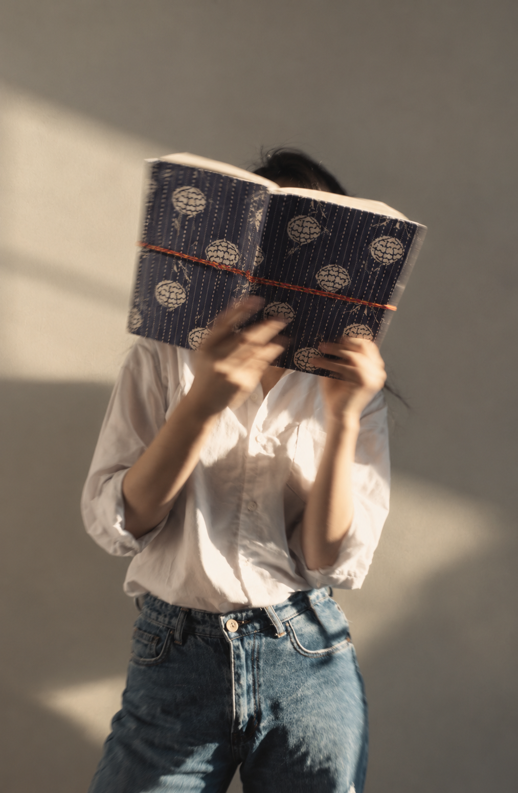 Navy Geometric block print fabric diary held open in front of the face, white shirt and jeans, sunlight on a grey wall