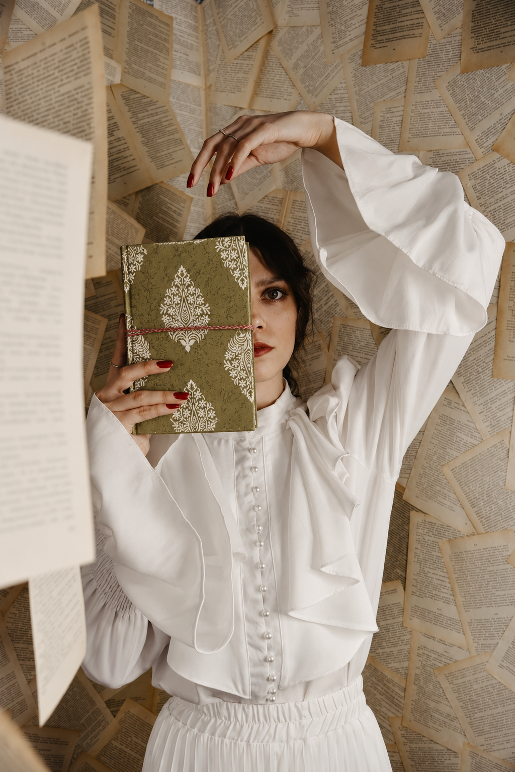 Olive Dabu block print fabric diary held beside the face against a wall of printed book pages, white ruffled blouse
