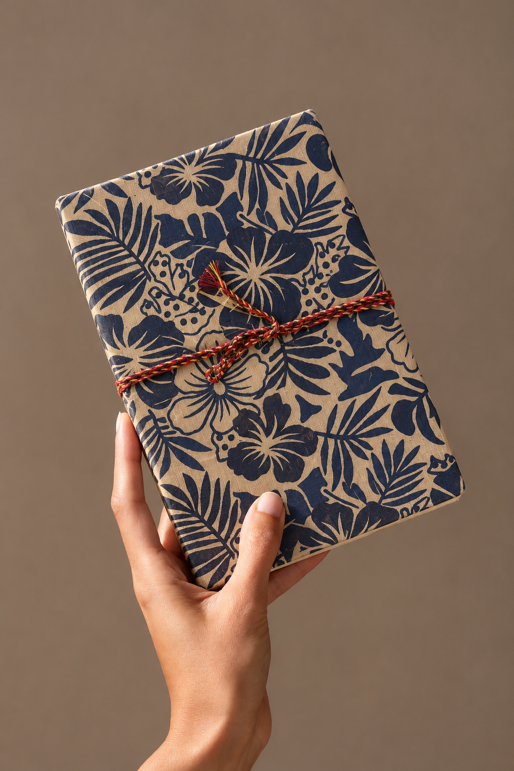 Indigo Tropics block print fabric diary raised in one hand against a taupe backdrop, braided cord tied in a bow