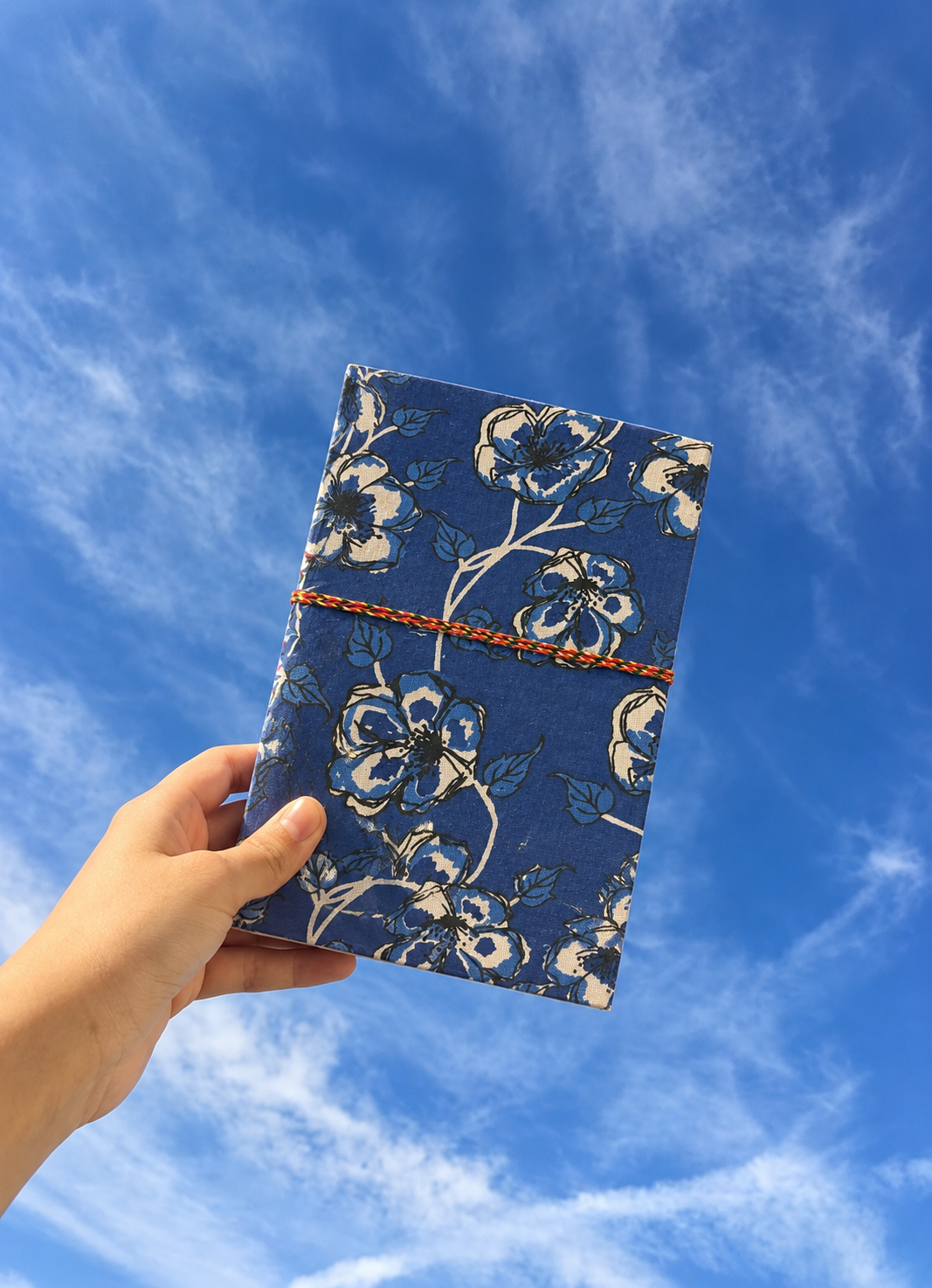 Neela Phool block print fabric diary held up in one hand against a blue sky with wispy clouds