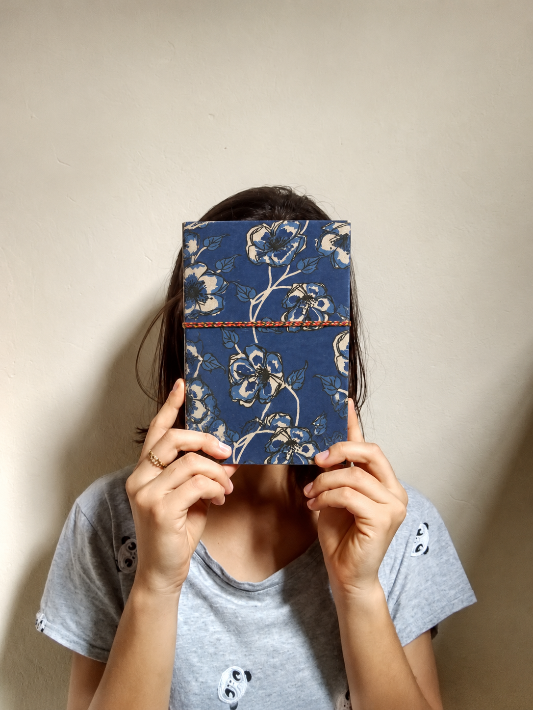Neela Phool block print fabric diary held up in front of the face in both hands, grey T-shirt, beige wall