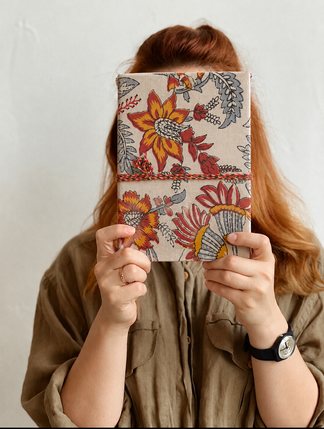 Hara Mahal block print fabric diary held up in front of the face, khaki shirt and a black watch, white wall