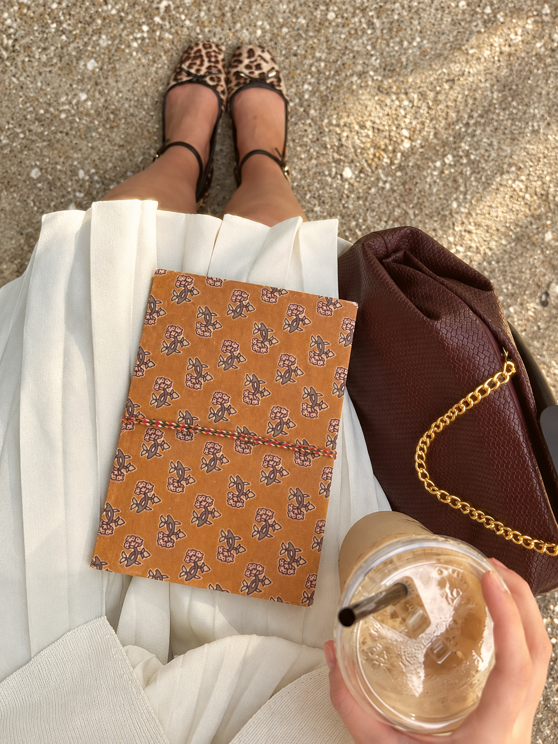 Amber Buti block print fabric diary on a white pleated skirt, seen from above with iced coffee and a burgundy chain bag