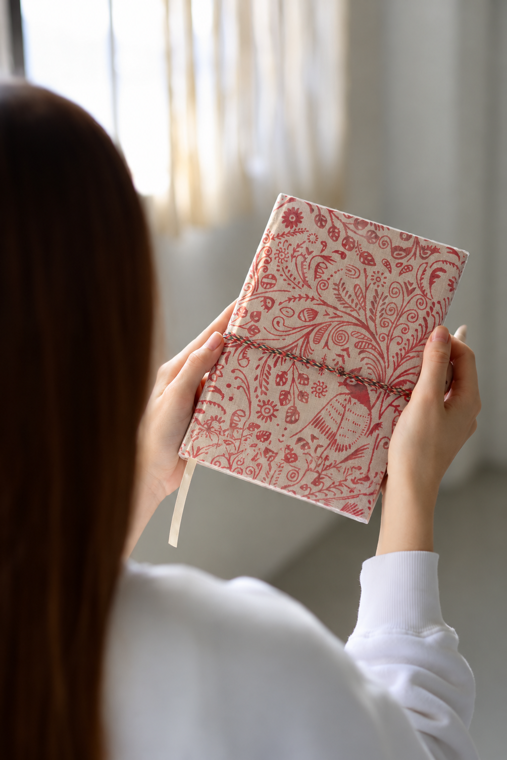 Crimson Peacock block print fabric diary held up by a woman seen from behind, near a window with sheer curtains