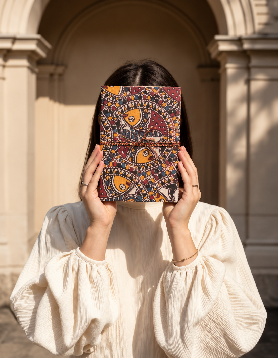Madhubani Carnival block print fabric diary held up to hide the face under a stone archway, cream puff-sleeve blouse