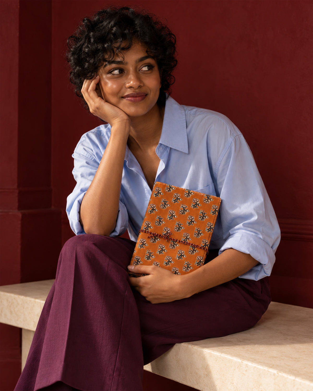 Amber Buti Block Print Fabric Diary, 200 Unruled Pages, held on the lap, blue shirt and aubergine trousers, maroon wall
