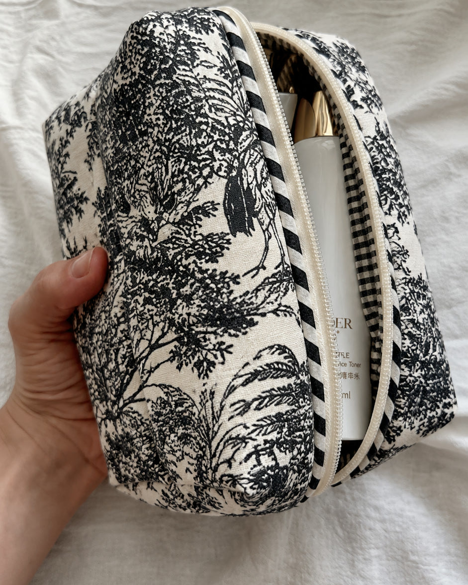 Hand holding the large Angan toile pouch, unzipped with a skincare bottle inside, showing the black-and-white striped zip trim