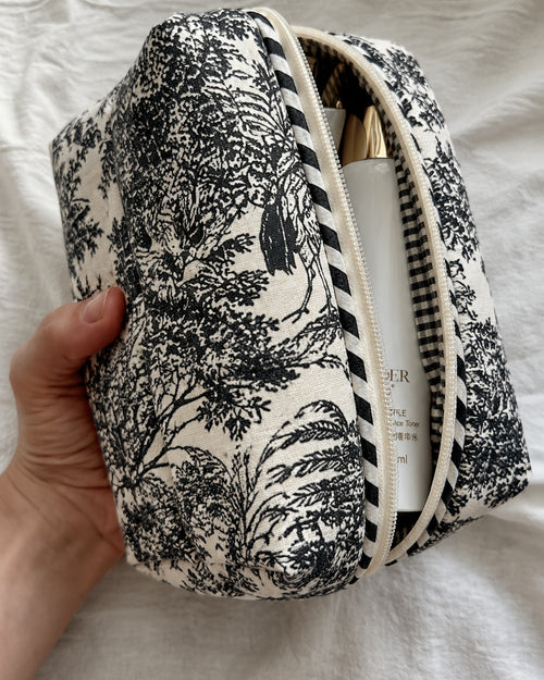 Hand holding the large Angan toile pouch, unzipped with a skincare bottle inside, showing the black-and-white striped zip trim