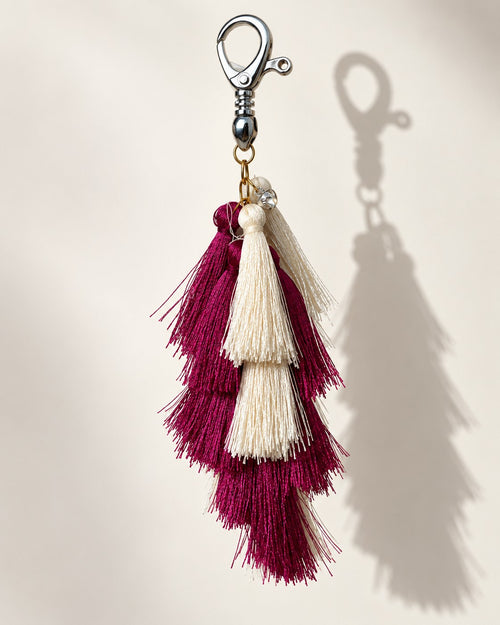 Aabha silk tassel bag charm: a small clear bead at the head, then long tiers of berry silk with ivory tassels set between them. It hangs from a silver-tone swivel clasp.