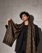 Shahi black Ajrakh modal silk stole held open over a black blazer against a concrete wall