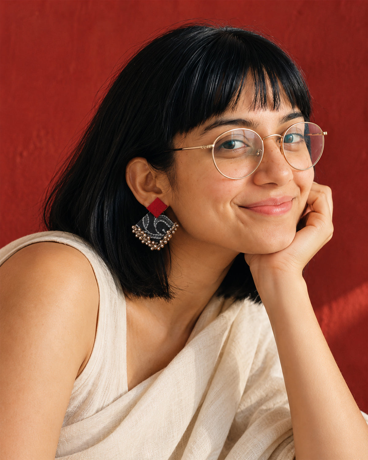 Black & Maroon Geometric Ghungroo Earrings, worn, head resting on the hand, round glasses, white top, vermilion red wall