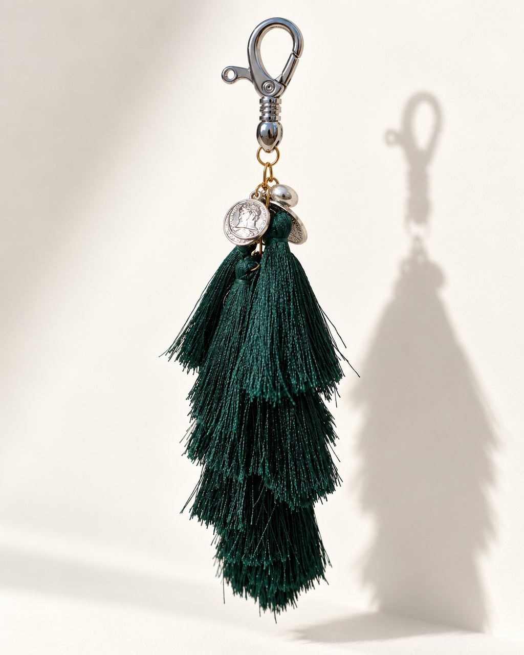 Neel Van silk tassel bag charm: embossed silver-tone coins and a small clear bead at the head, above a cluster of tiered forest-green silk tassels. It hangs from a silver-tone swivel clasp.