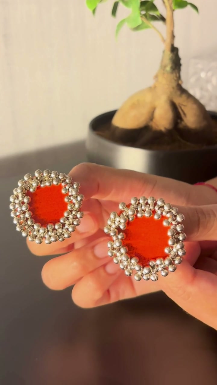 Orange Ghungroo Stud Earrings: Orange Ghungroo Stud Earrings held between the fingers and turned in the light: orange cotton centres ringed with two rows of ghungroo bells