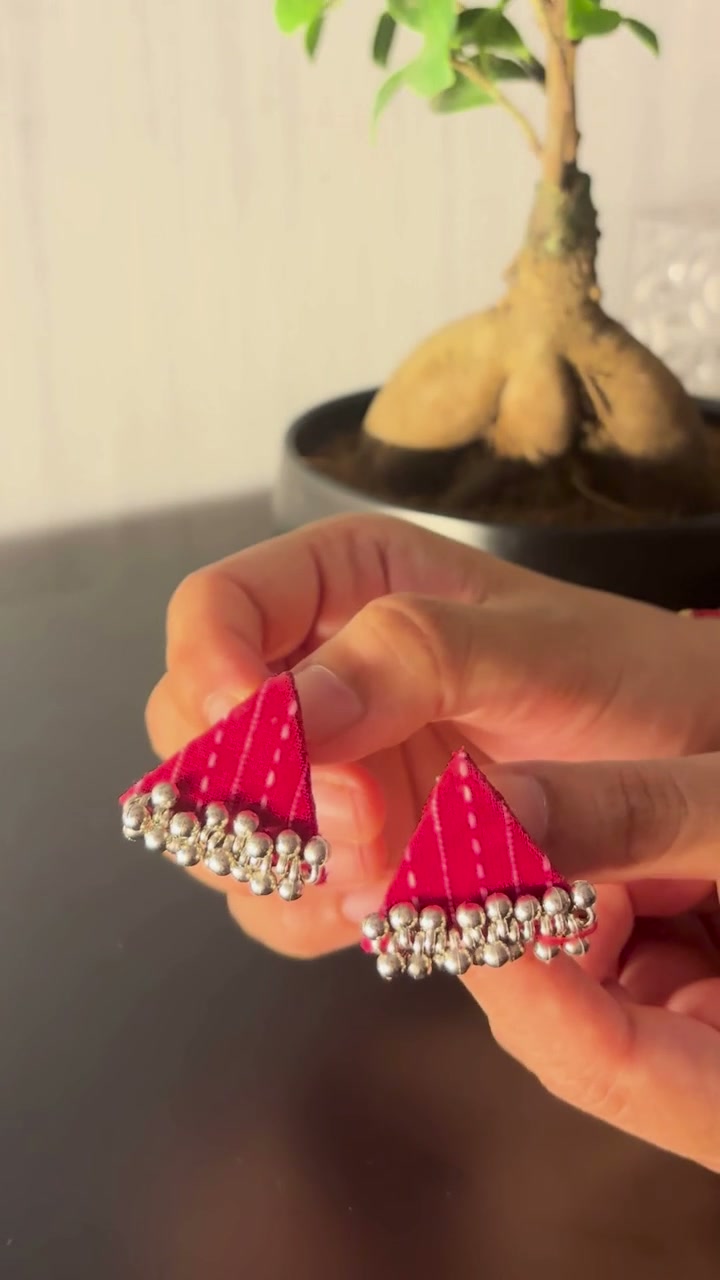 Riwaayat Pink Stitch Triangle Earrings: Riwaayat Pink Stitch triangle earrings held between the fingers: rani pink fabric lined with white running stitches and a row of ghungroo bells