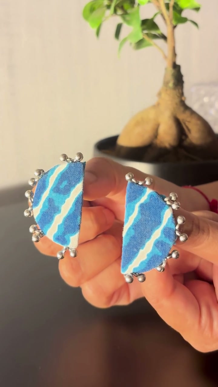 Leher Sky Blue Half Moon Earrings: Leher Sky Blue half moon earrings held between the fingers: sky blue and white lehariya stripes edged with silver-tone ghungroo beads
