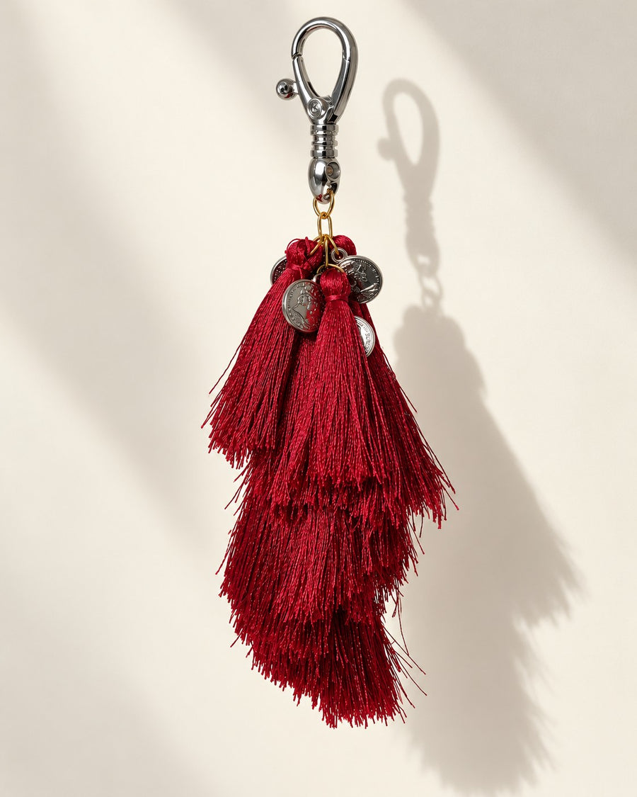 Raktima silk tassel bag charm: a cluster of embossed silver-tone coins at the head, above tiered ruby red silk tassels. It hangs from a silver-tone swivel clasp.