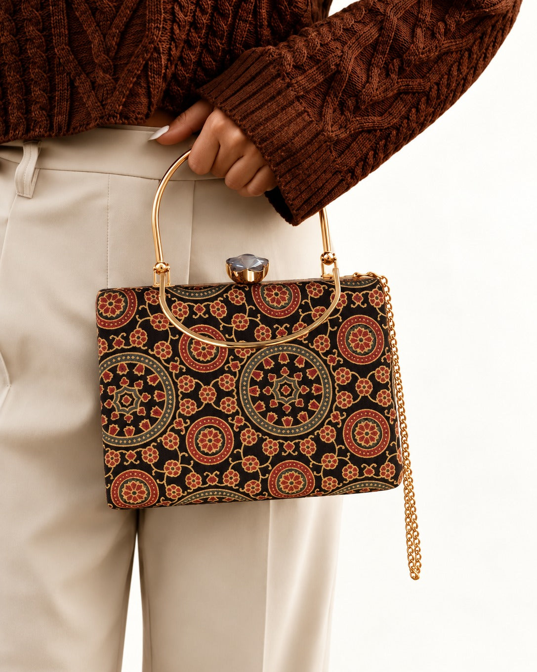 Ruhani black medallion-print Mashru silk handbag carried by its top handle against ivory trousers and a brown knit