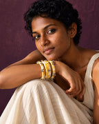 Lariya Yellow Leheriya Fabric Bangles with Ghungroo, Kada Set of 3, worn on the wrist, arms folded over the knees, white outfit, aubergine wall