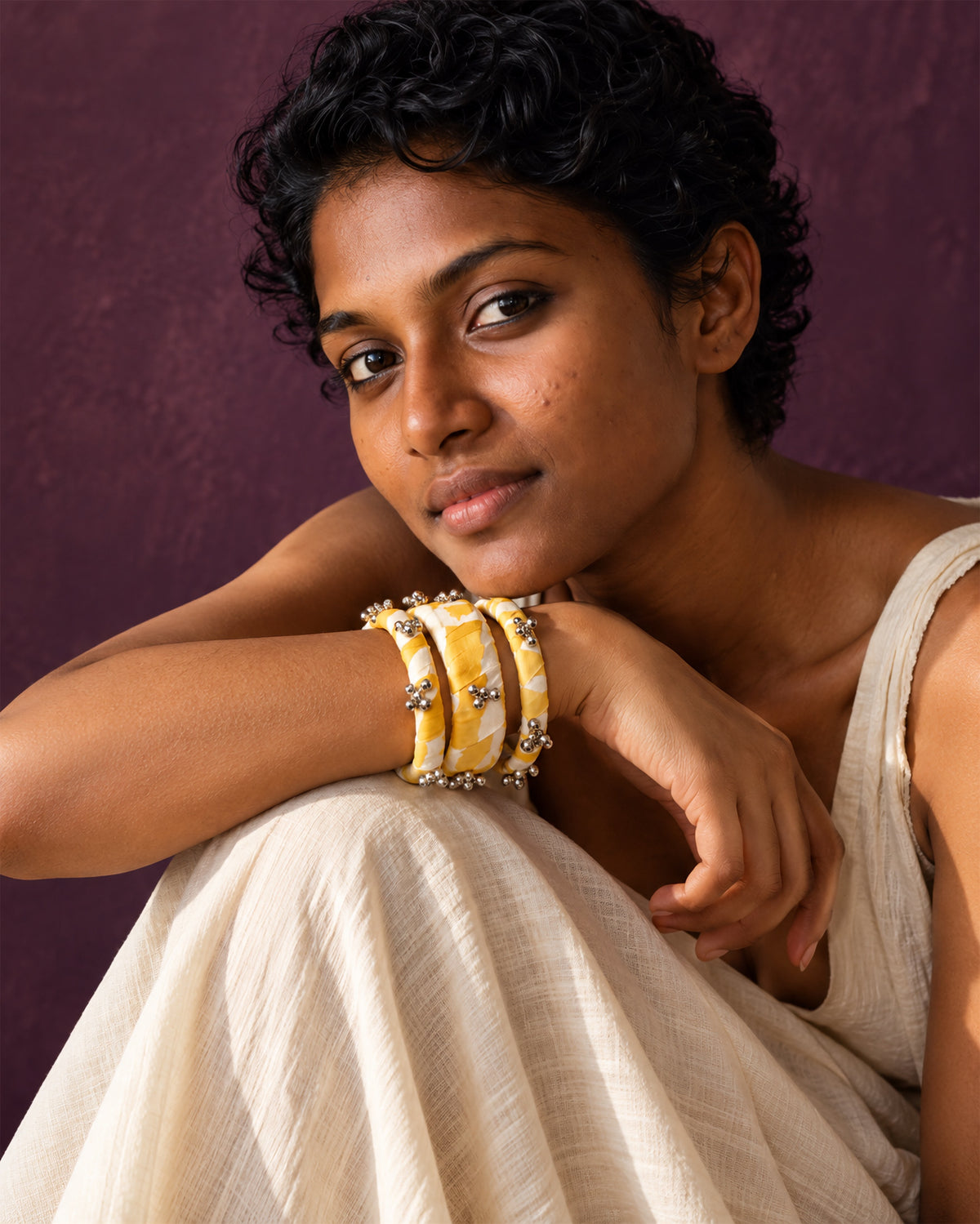 Lariya Yellow Leheriya Fabric Bangles with Ghungroo, Kada Set of 3, worn on the wrist, arms folded over the knees, white outfit, aubergine wall